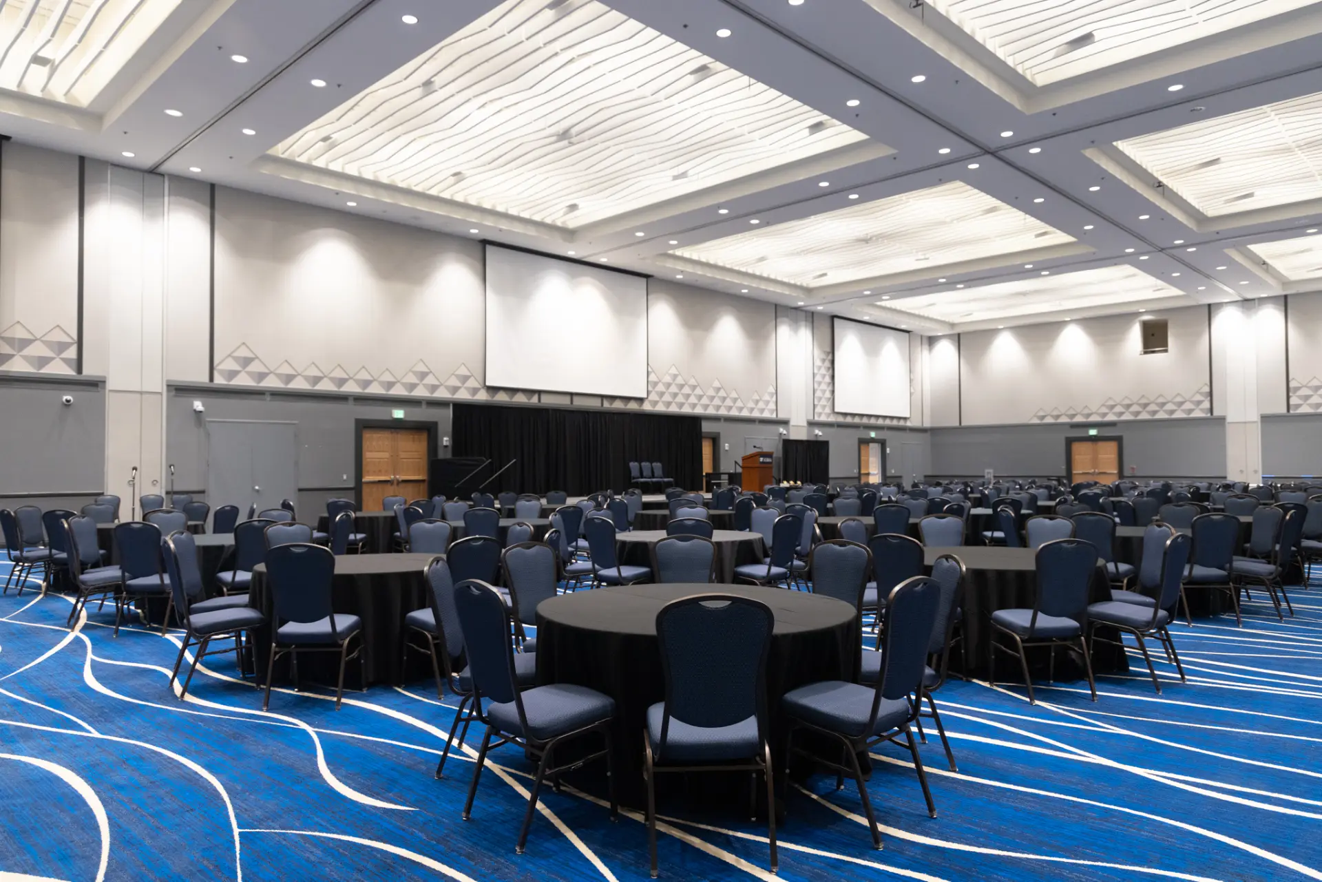 A large conference room with a blue carpet featuring white wavy lines, a high ceiling with recessed lighting, and a row of tables surrounded by chairs.