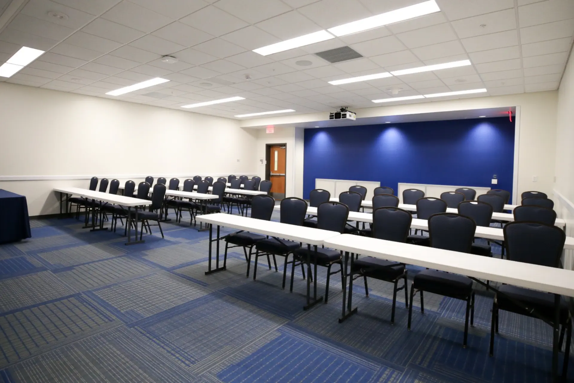 An empty conference room with a long table and chairs, featuring blue and gray carpeting, a blue wall, and a projector screen.