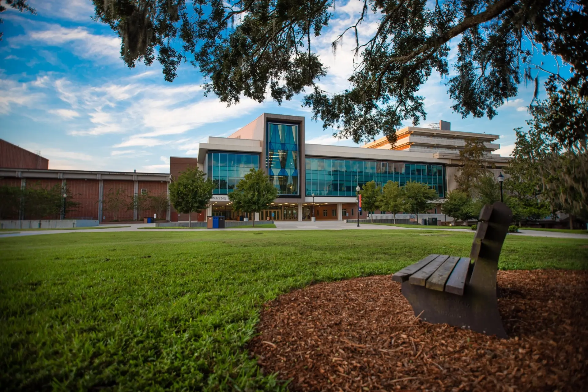 A large building with a glass facade, surrounded by trees and greenery, featuring a bench in the foreground.