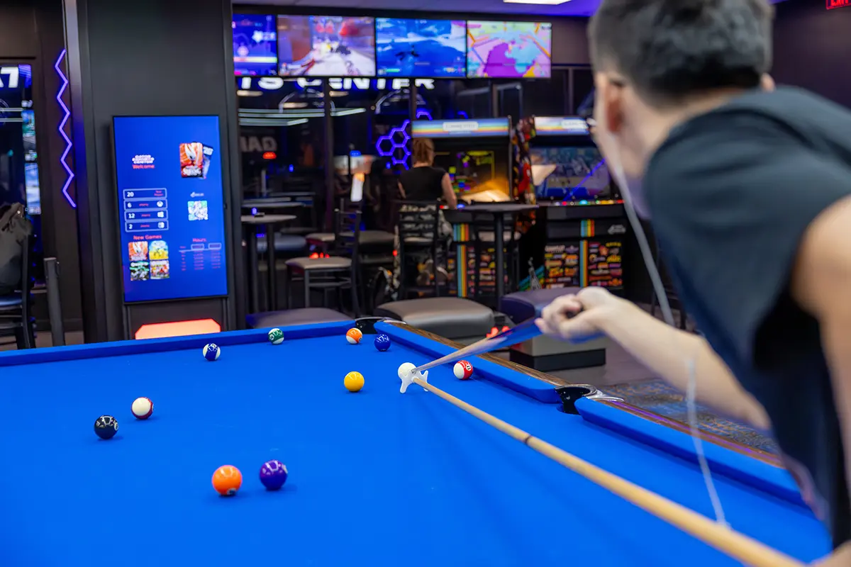 A person in a black shirt plays pool in a game room, with a blue table and balls scattered about.